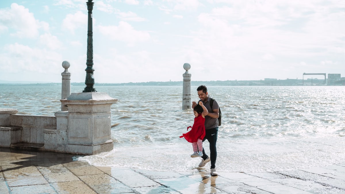 A father joyfully swings his daughter by the sea on a bright day, capturing a playful moment.
